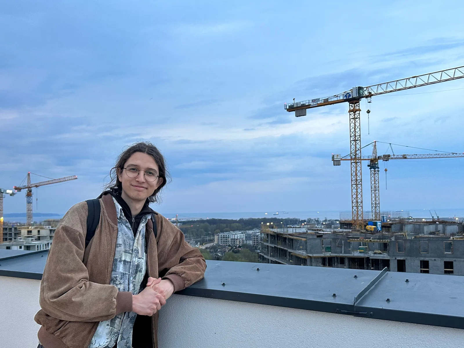 Patryk uśmiechnięty, oparty o barierkę na dachu, w tle panorama miasta z dźwigami i morzem
