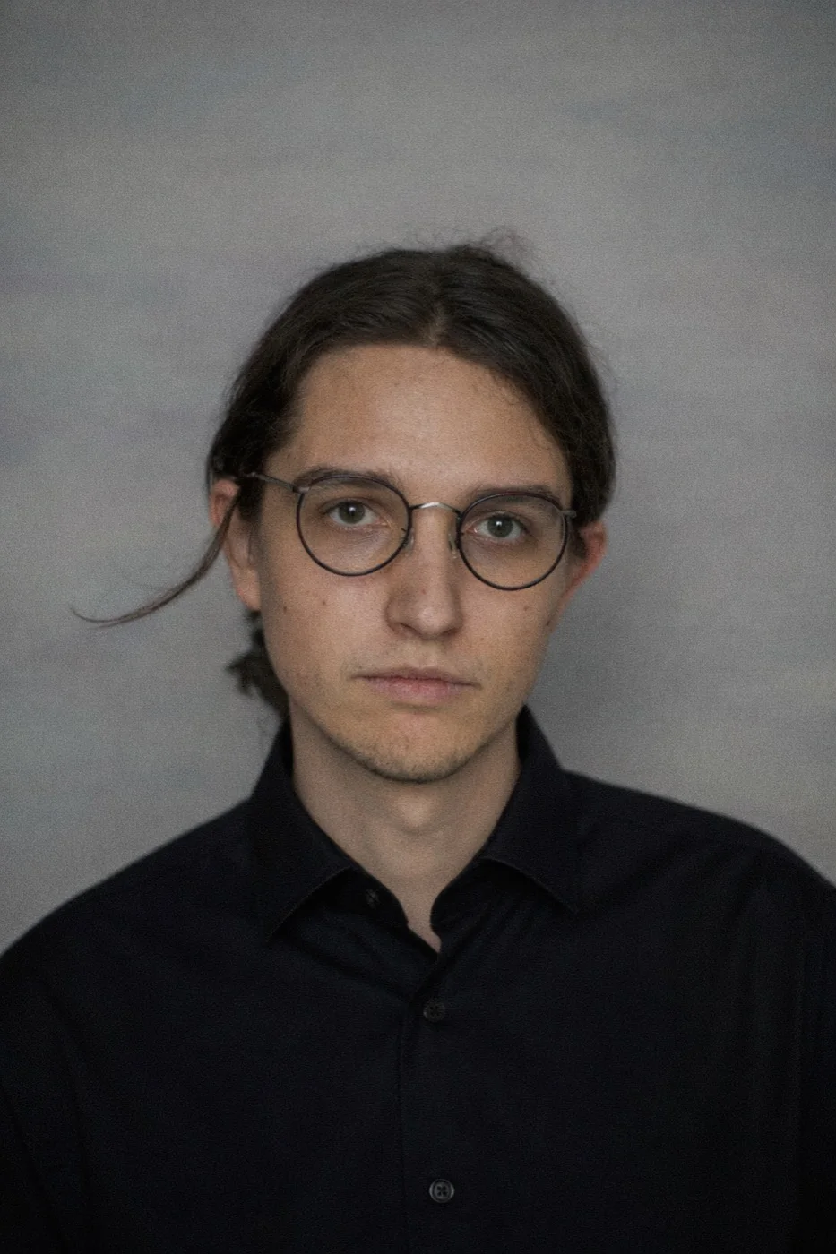 Patryk Mikołajczyk, portret twarzą do obiektywu, w ciemnej koszuli i okrągłych okularach, na szarym tle