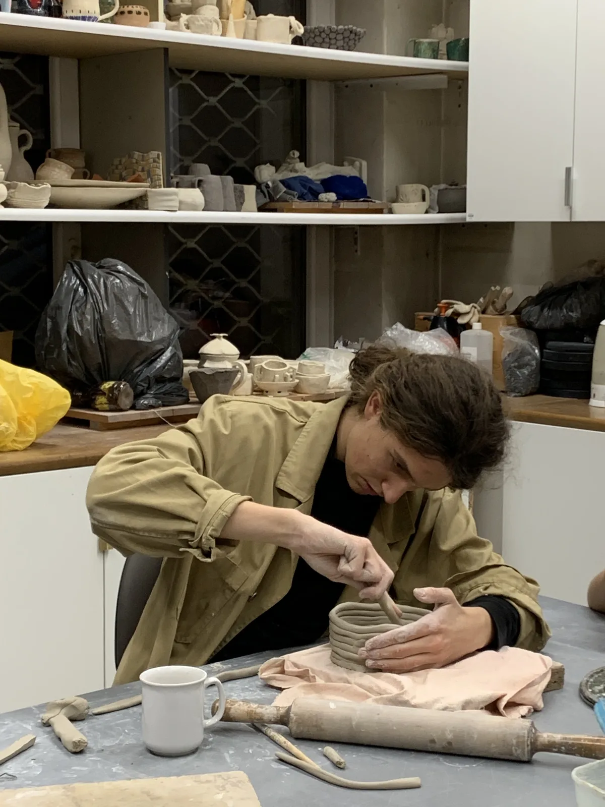 Patryk w pracowni ceramicznej, skupiony lepi z gliny naczynie metodą wałeczkową, w tle półki z ceramiką