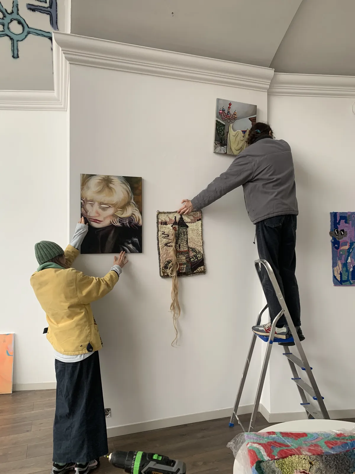 Dwie osoby rozwieszają prace na białej ścianie galerii, jedna poprawia malarski portret, druga na drabinie wiesza tkaninę