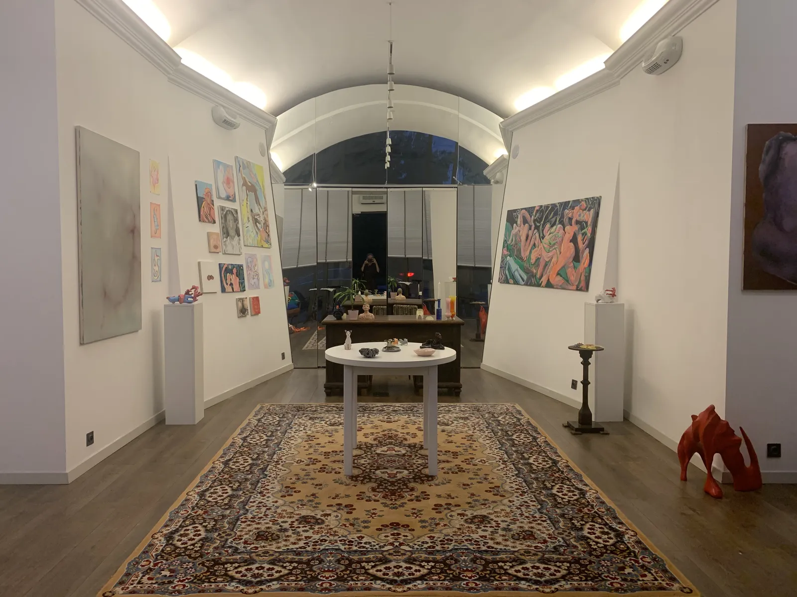 Wnętrze galerii, białe ściany z obrazami, pośrodku okrągły stół na dywanie, z tyłu łukowe okno